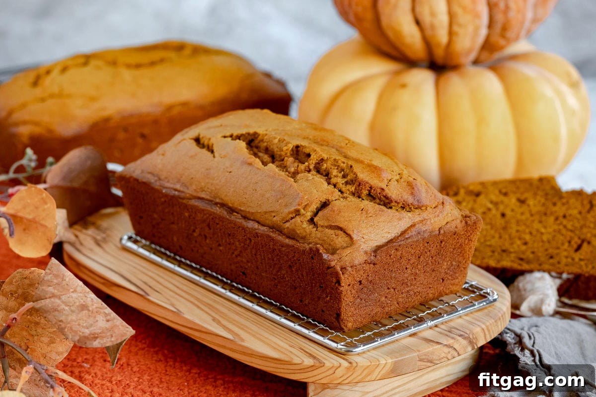 A whole loaf of pumpkin bread prominently displayed on an olive wood board, with another loaf softly blurred in the background.