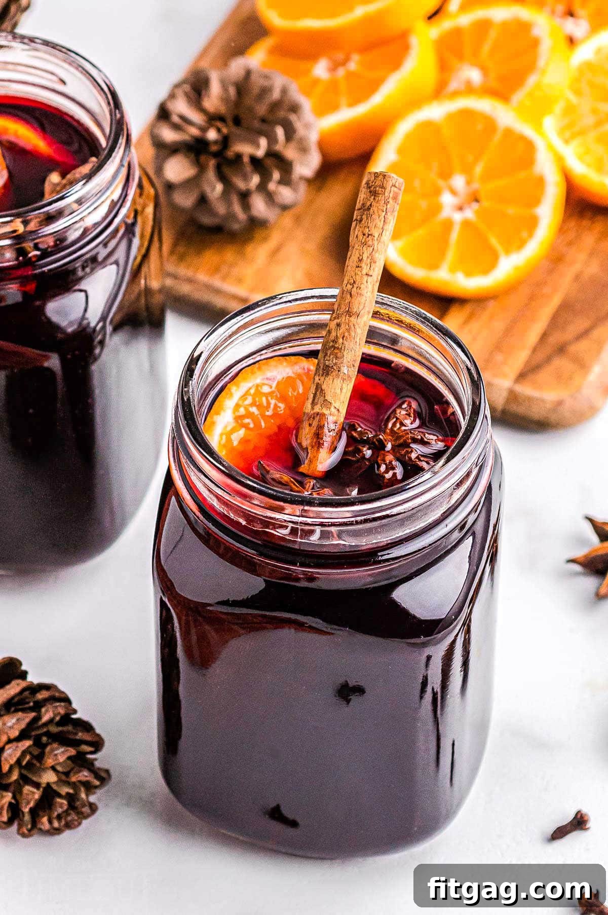 A glass mug filled with richly spiced mulled wine, beautifully garnished with a cinnamon stick, a vibrant orange slice, and an elegant star anise. Sliced oranges rest softly in the background, adding to the festive charm of the scene.