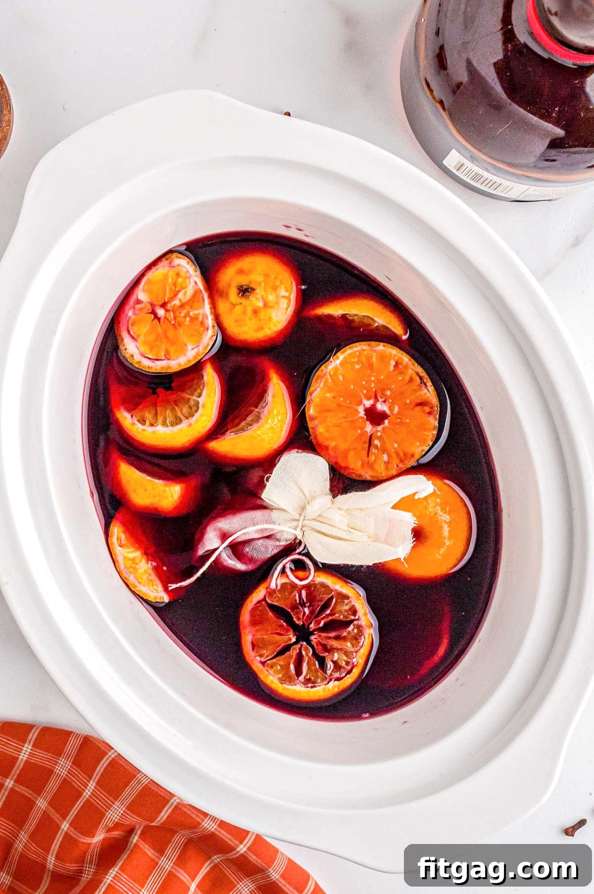A small spice satchel, made of cheesecloth and tied with string, rests gently amidst the rich red wine and sliced oranges within a white crockpot. This image perfectly captures the initial stage of infusing mulled wine with aromatic spices.