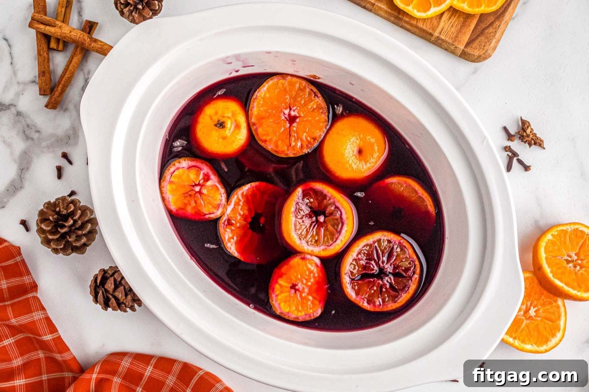 A top-down perspective captures the inviting sight of mulled wine simmering gently in a white crockpot, adorned with bright orange slices and warm cinnamon sticks, ready for serving.