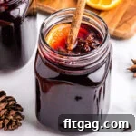 A glass jar filled with rich spiced wine, elegantly topped with a cinnamon stick, a vibrant orange slice, and star anise, with softly sliced oranges blurred in the background, creating a warm and inviting scene.