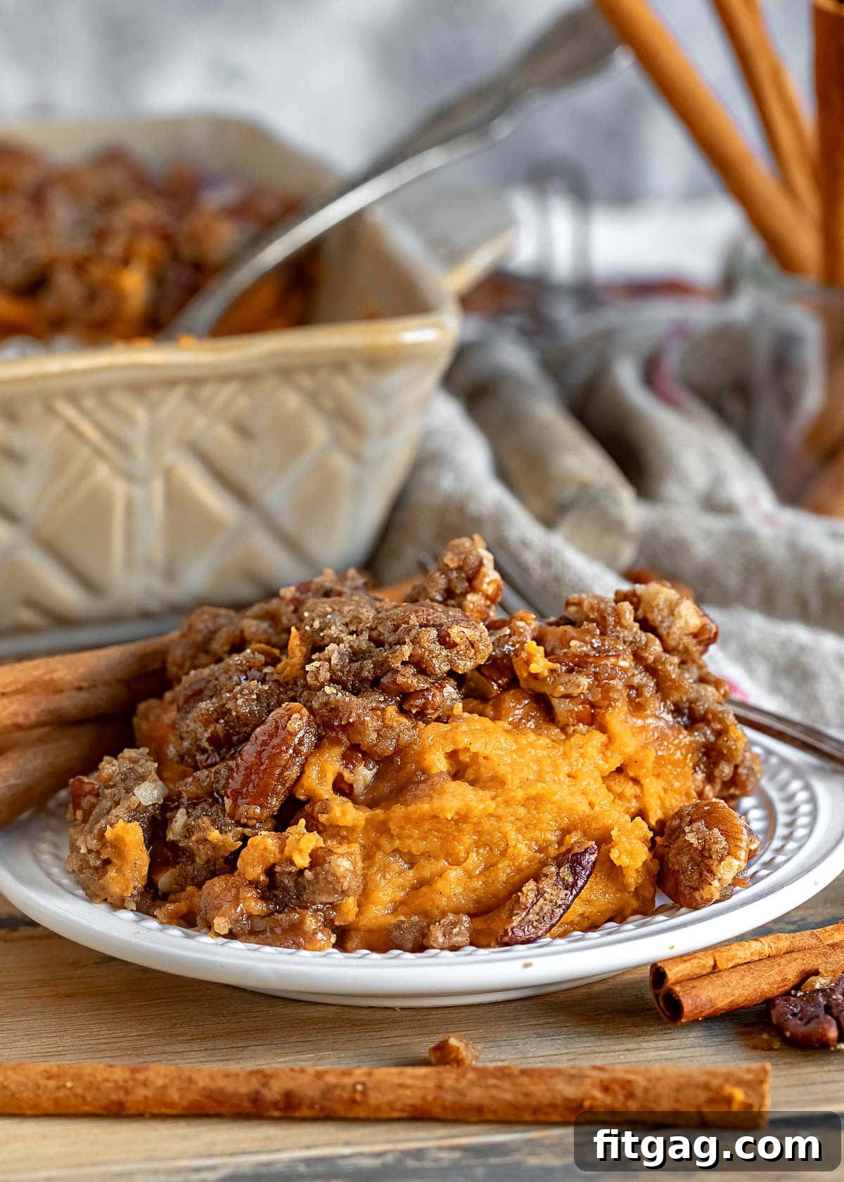 Small plate featuring a serving of golden-brown sweet potato casserole with a pecan streusel topping. A larger baking dish with the remaining casserole sits in the background.