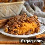 Small plate with a serving of sweet potato casserole on it. Baking dish with the rest of the casserole in background. casserole is topped with a pecan streusel.