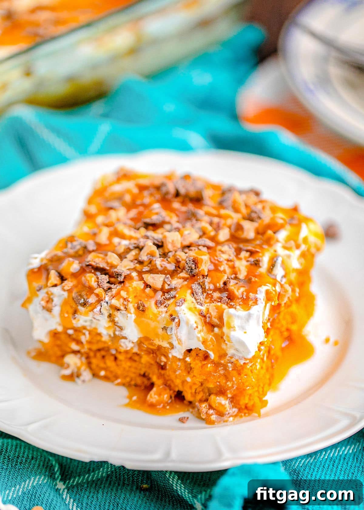A slice of Better Than Anything Pumpkin Cake on a white plate with a bright blue napkin in the background. The cake is topped with cool whip and Heath toffee bits.