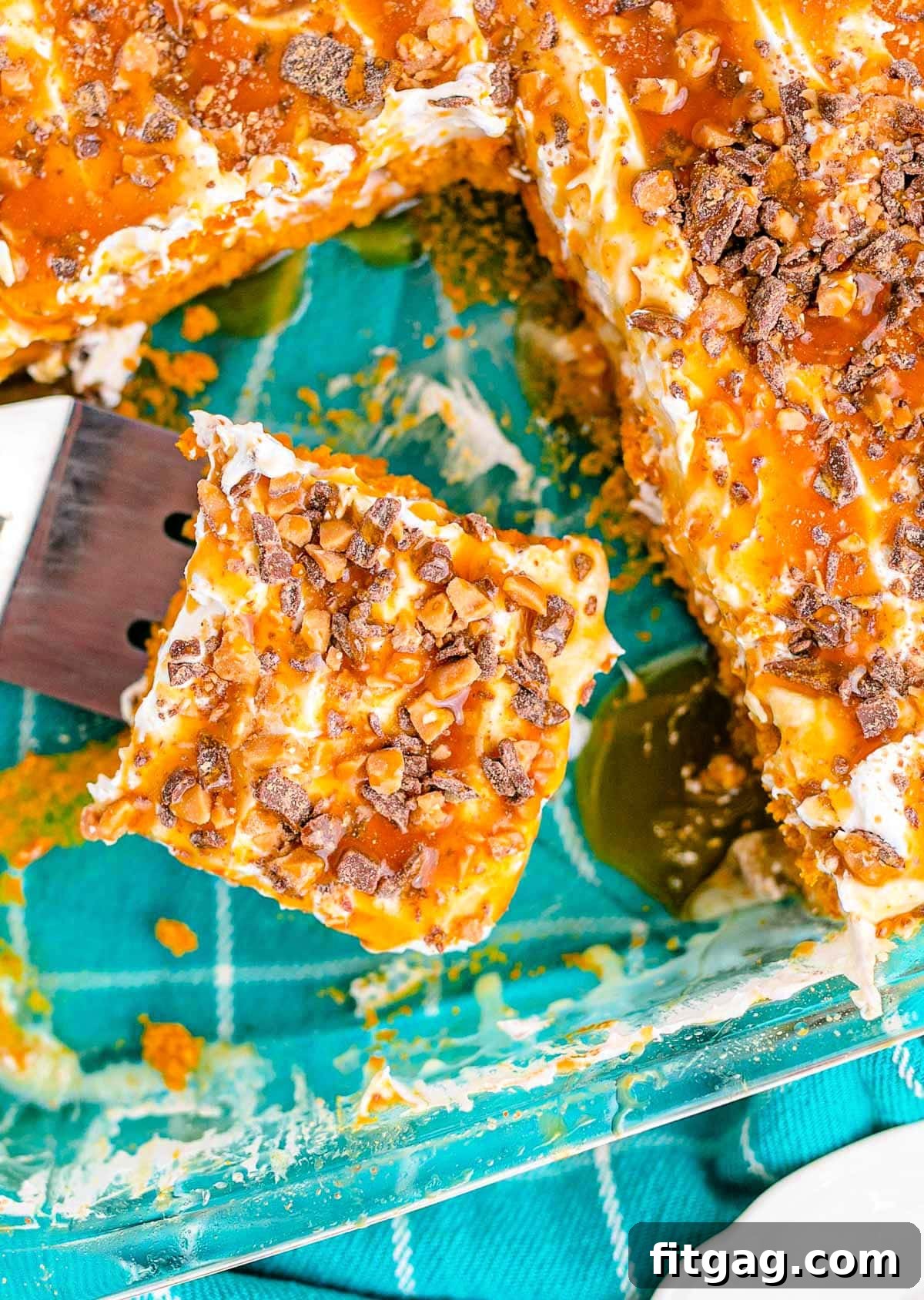 Top-down view of a pumpkin poke cake topped with cool whip, caramel, and toffee bits. Several pieces have been removed, and one piece is on a serving utensil.