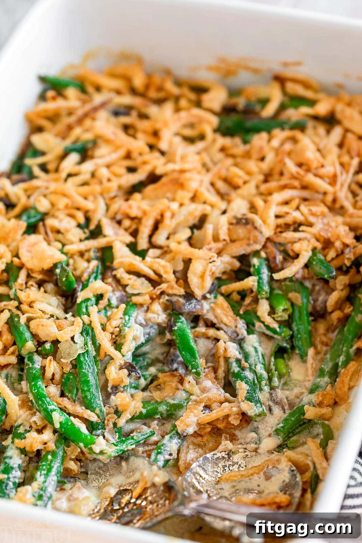Crispy Onion Green Bean Delight 2 A large white baking dish filled with homemade green bean casserole, generously topped with golden-brown fried onions. Two silver serving spoons are nestled within the dish, indicating a serving or two has been removed, showcasing the creamy interior.