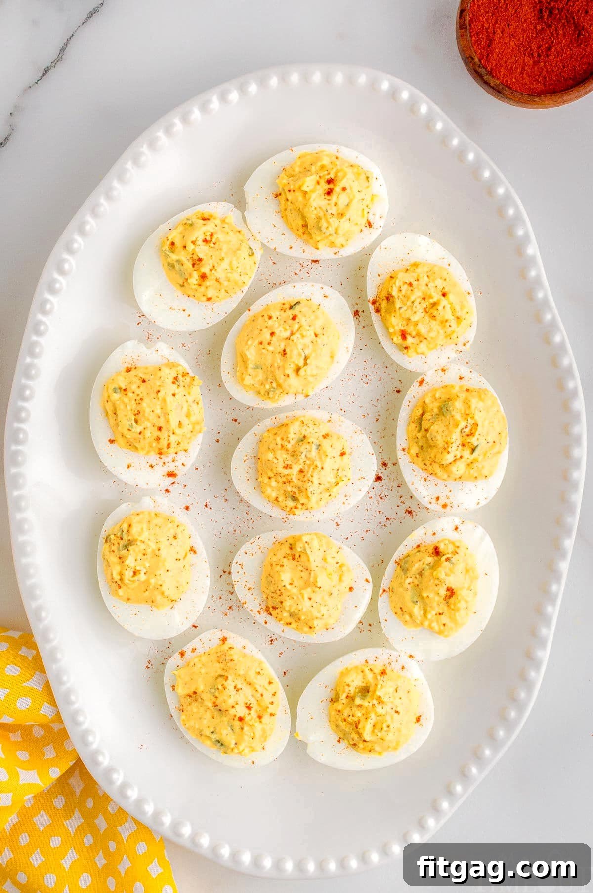 A pristine white serving tray generously filled with a dozen classic deviled eggs, artfully garnished with a sprinkle of vibrant paprika. A soft yellow napkin peeks from the corner, adding a touch of warmth to the inviting presentation.