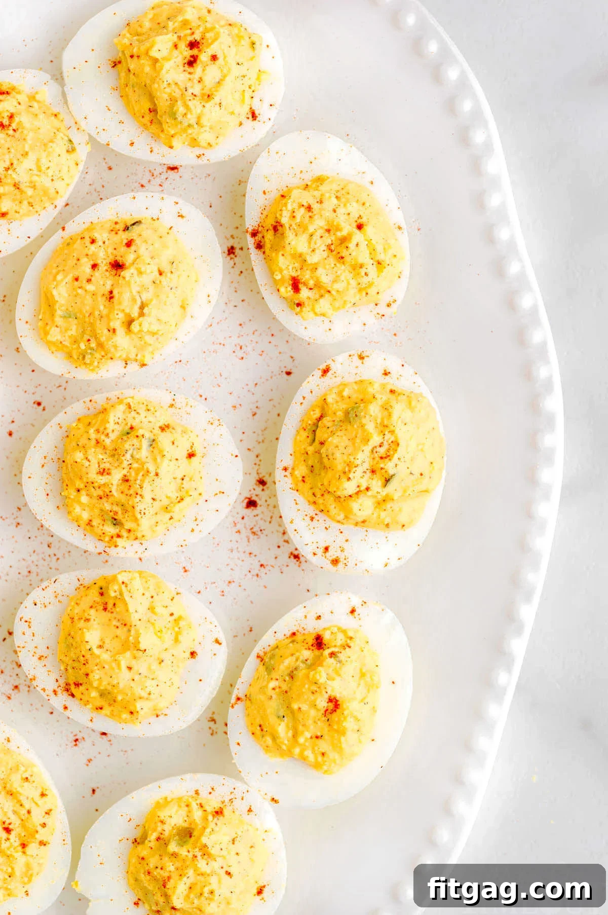 A top-down view of a white platter, half-filled with classic deviled eggs, each perfectly garnished with a dusting of vibrant red paprika, creating an inviting and artful presentation.
