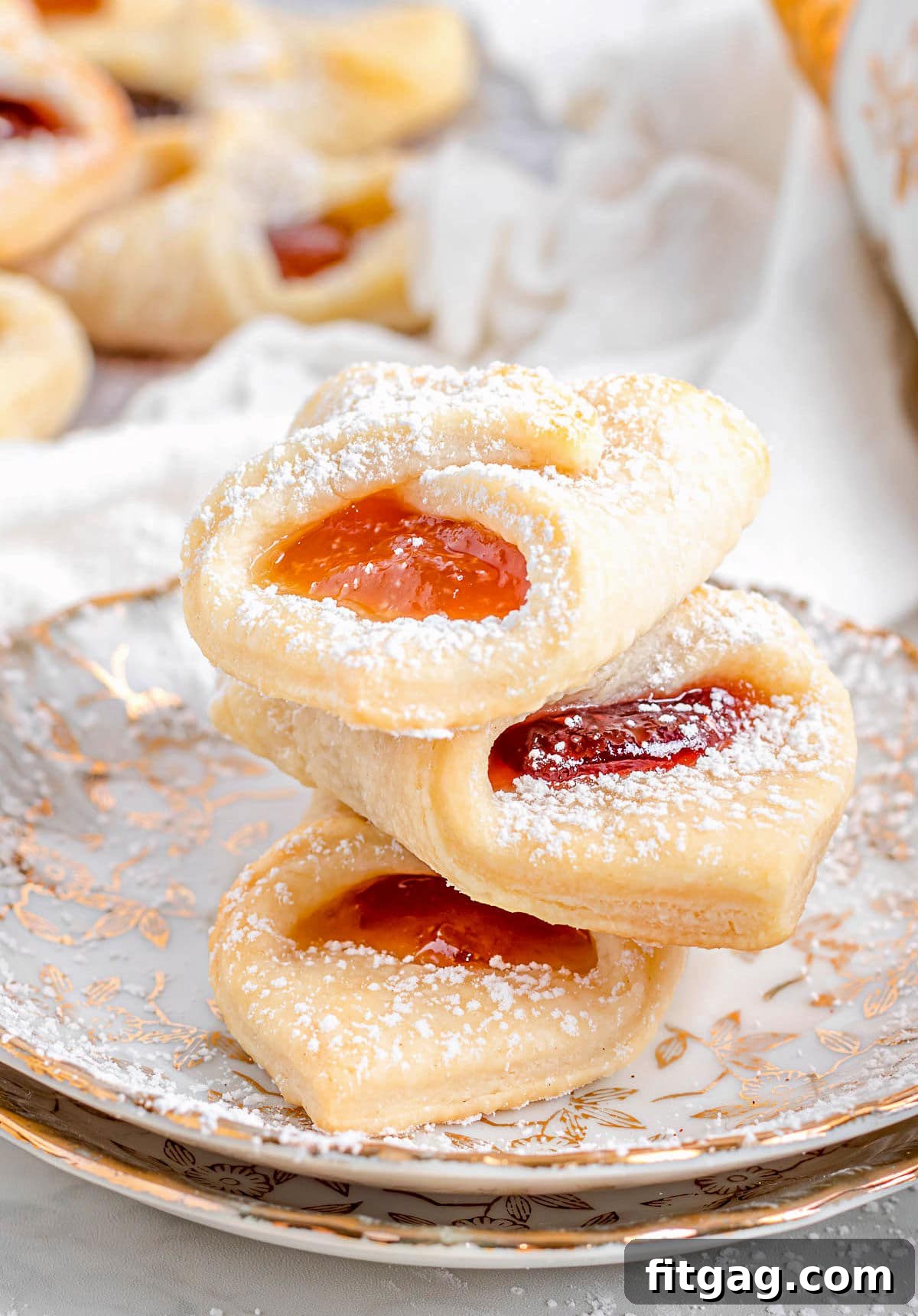 Three Kolaczki cookies stacked on two gold-rimmed plates, showcasing their powdered sugar and jam filling.