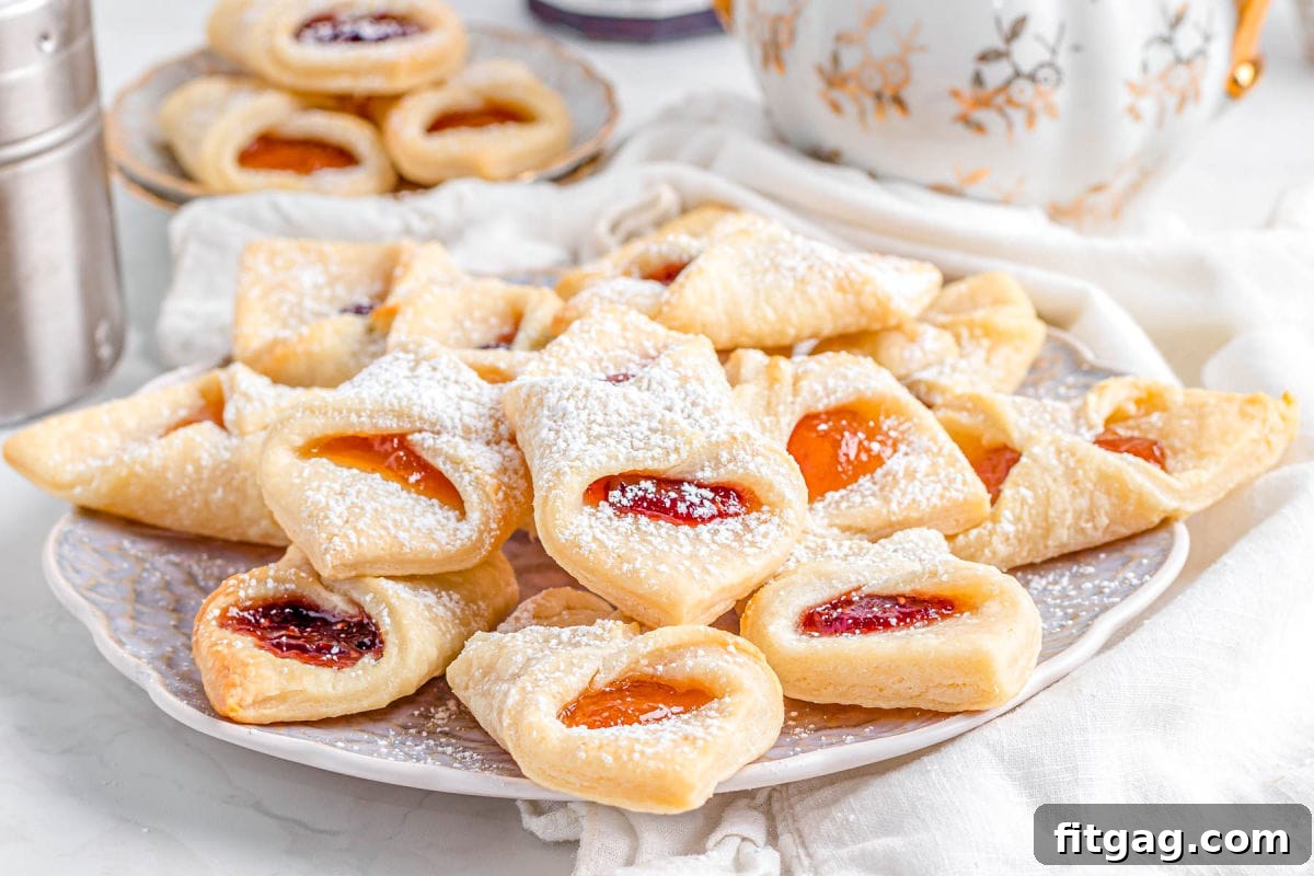 A large china plate generously stacked with Kolachy cookies, beautifully dusted with powdered sugar and showcasing their vibrant jam centers, ready to be enjoyed.