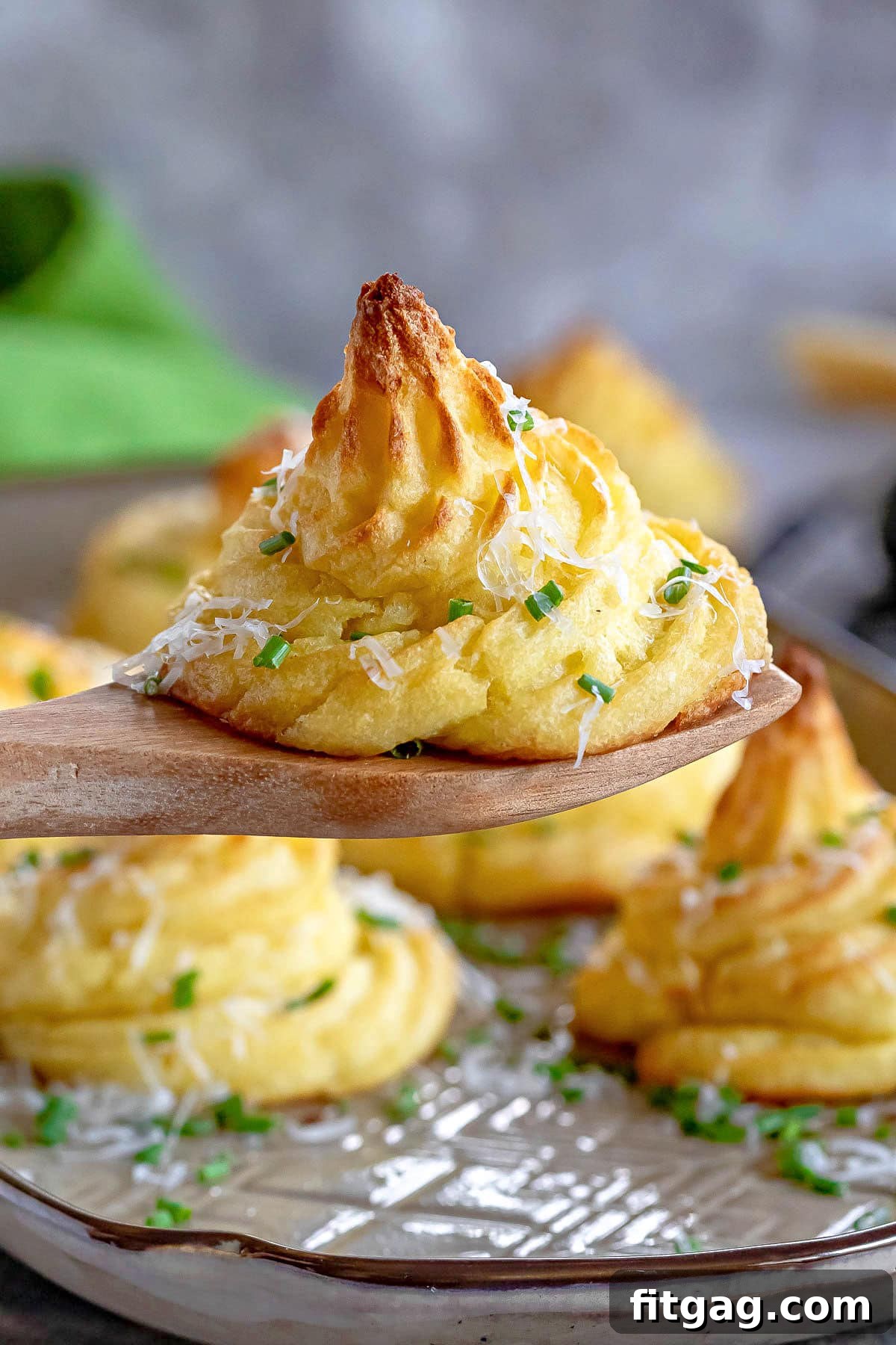 A single elegantly piped Duchess potato, garnished with fresh chives and grated Parmesan, is held aloft by a wooden serving spoon over a tray of other Duchess potatoes.