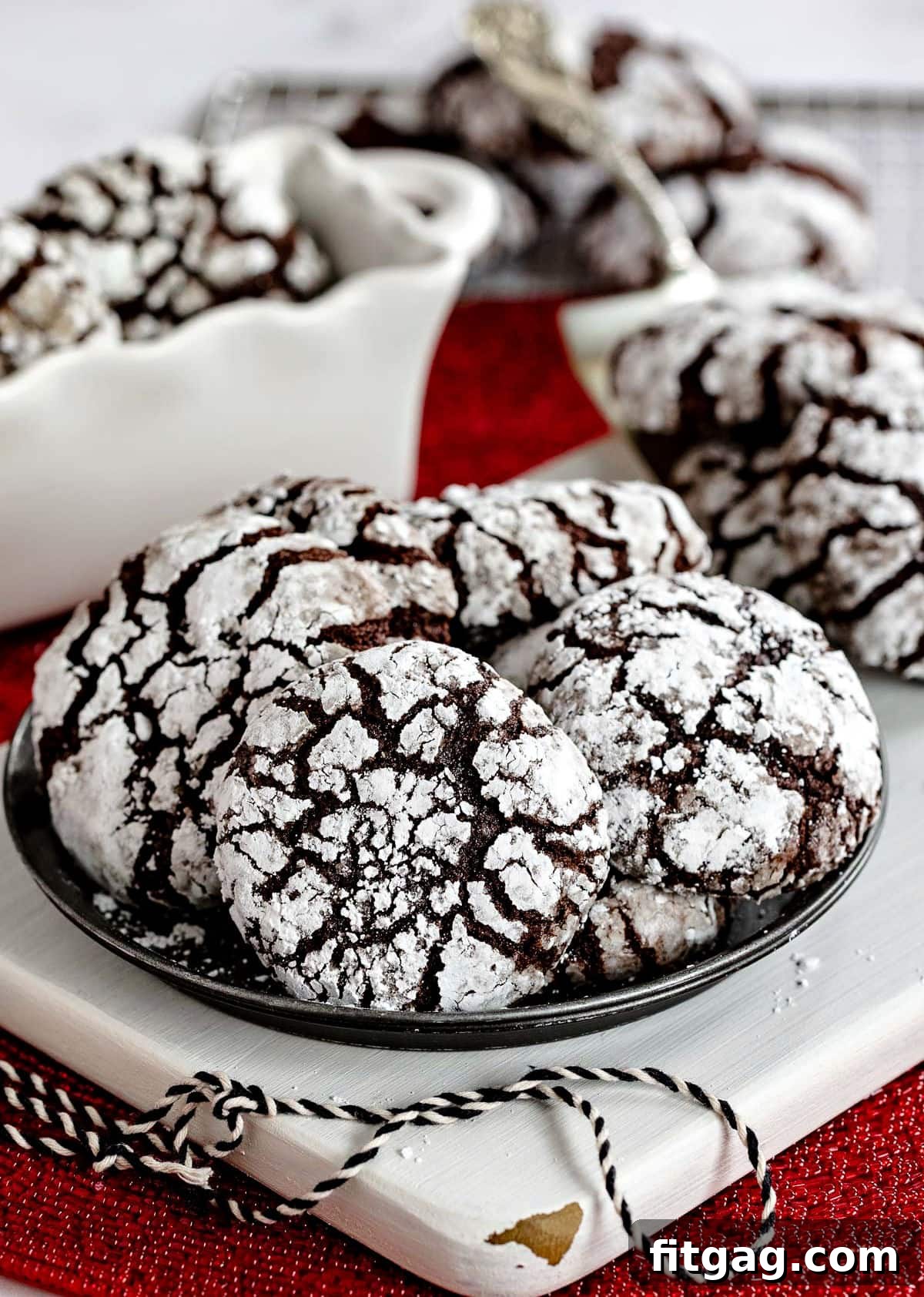 Small metal plate with six chocolate crinkle cookies sitting on a white wood board with more cookies on a server and cooling rack in background. These cookies are moist and fudgy, perfect for holidays.