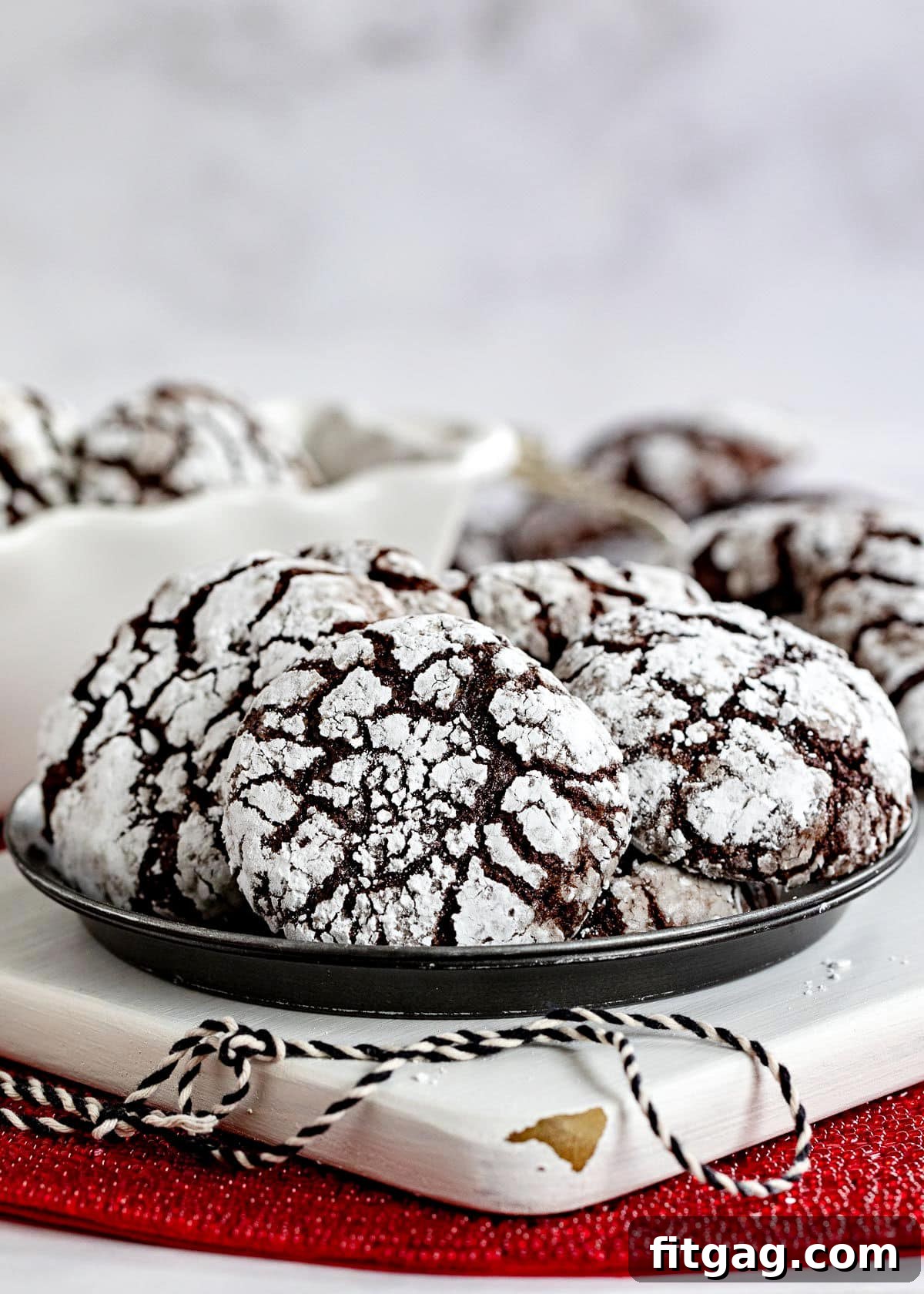 Plate of chocolate crinkle cookies sitting on white wood board on red sparkly placemat. Black and white twine sitting on board and more cookies can be seen in the background.