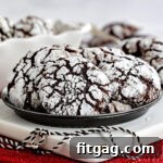 plate of chocolate crinkle cookies sitting on white wood board on red sparkly placemat. black and white twine sitting on board and more cookies can be seen in the background.