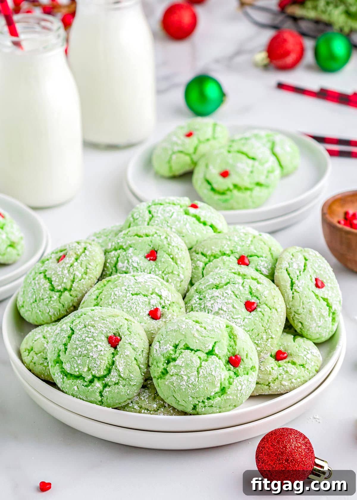 Grinchy Green Goodies 2 Vibrant green Grinch cake mix cookies adorned with bright red heart sprinkles, beautifully arranged on a white plate. In the background, festive Christmas ornaments and milk jars create a cheerful holiday scene.