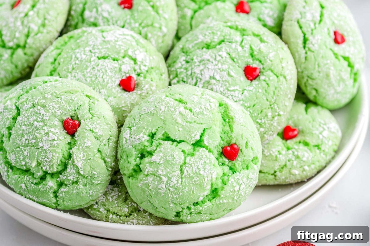 Grinchy Green Goodies 3 A charming stack of two white plates, laden with a generous pile of vibrant green Grinch cookies, each crowned with a delicate red heart sprinkle.