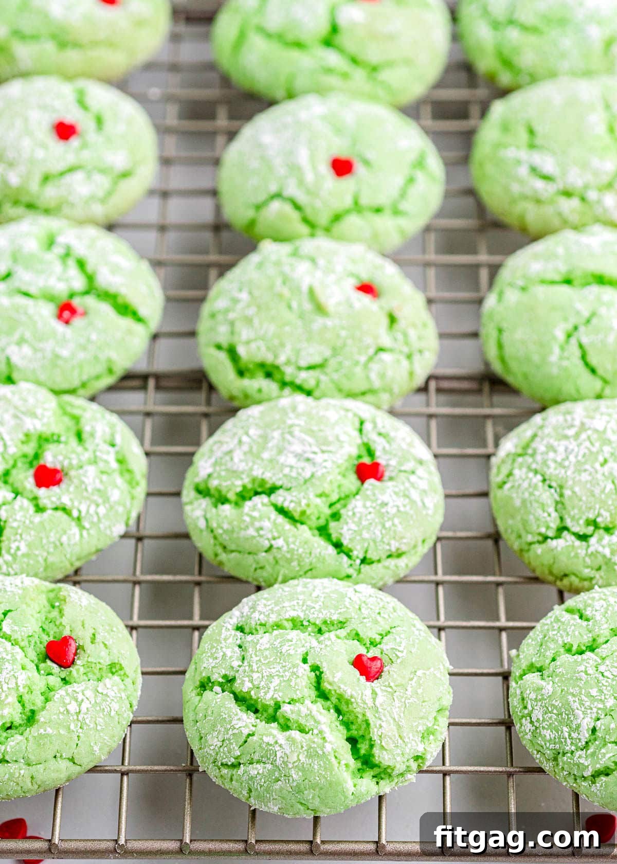 Grinchy Green Goodies 4 A neat row of vibrant green Grinch cookies, each topped with a single red heart sprinkle, cooling on a wire rack.