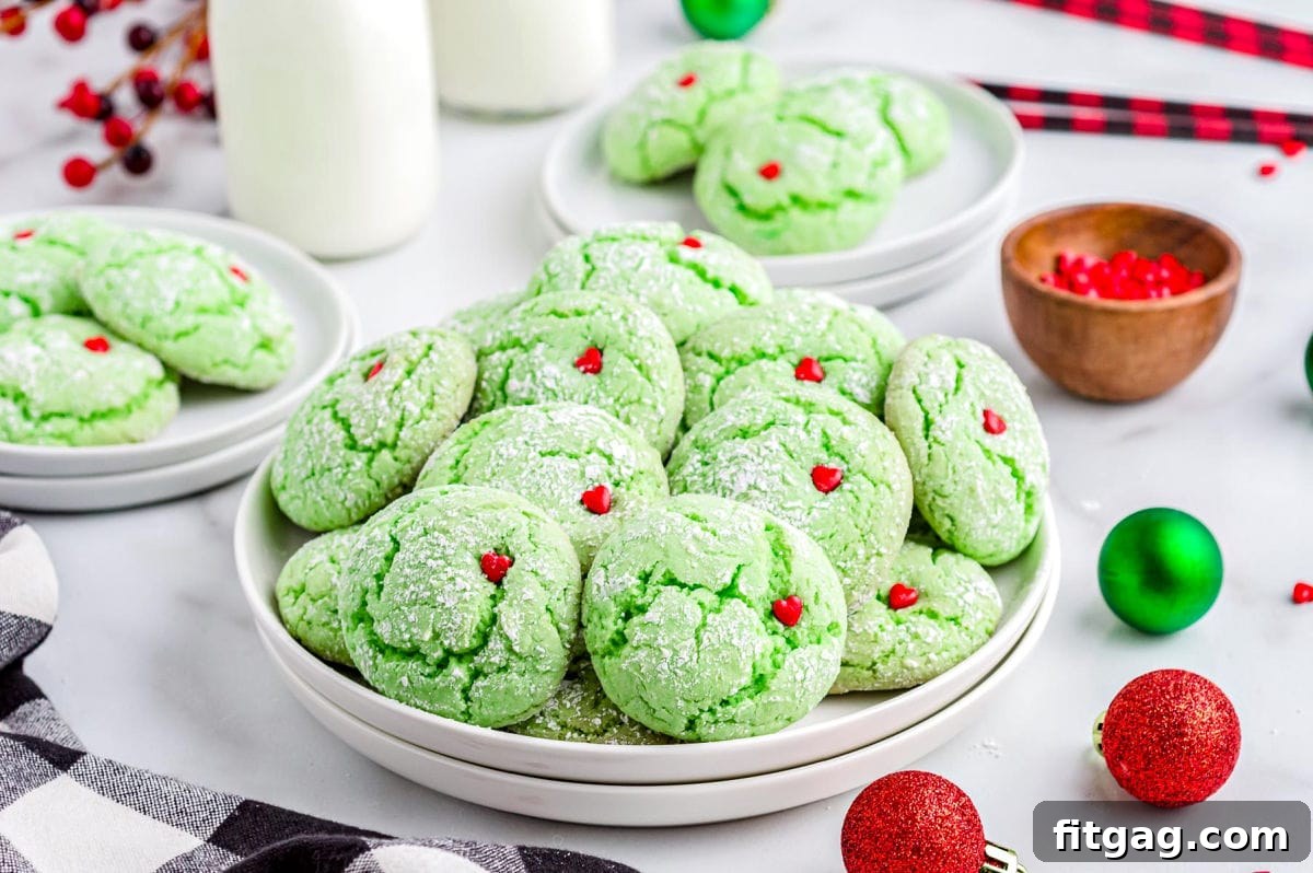 Grinchy Green Goodies 6 A festive display of green Grinch cake mix cookies with red heart sprinkles, arranged on a white plate with milk jars and scattered Christmas ornaments in the background.