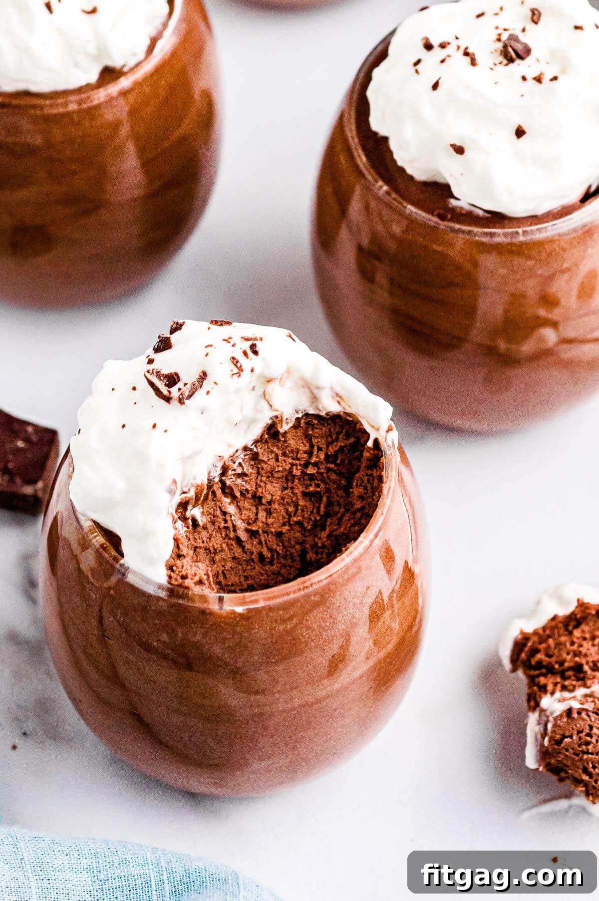 A wine glass filled with luscious chocolate mousse, revealing its airy, foamy texture after a spoonful has been taken. Two more glasses of mousse are blurred in the background.