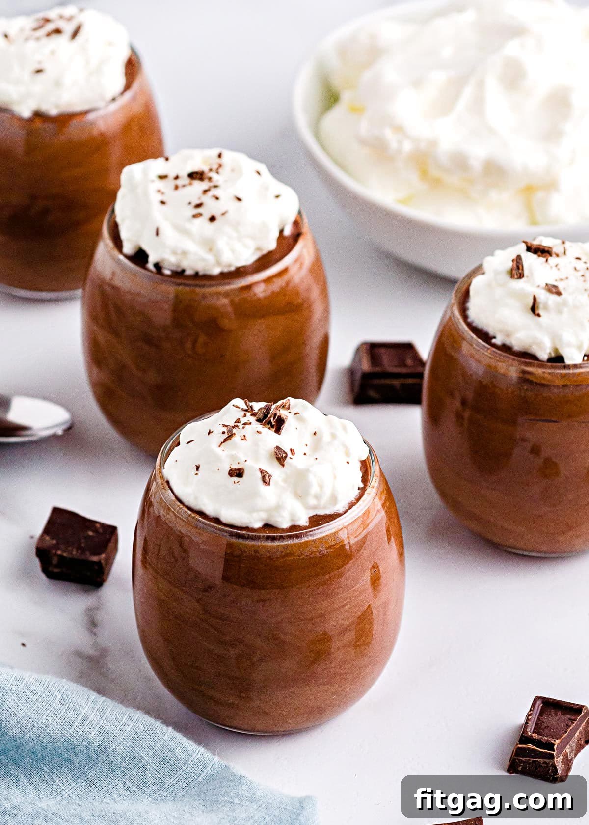 Four glasses filled with rich, dark chocolate mousse, each crowned with a generous swirl of whipped cream and a sprinkle of delicate chocolate shavings, positioned on a rustic wooden surface, ready to be enjoyed as a luxurious dessert.