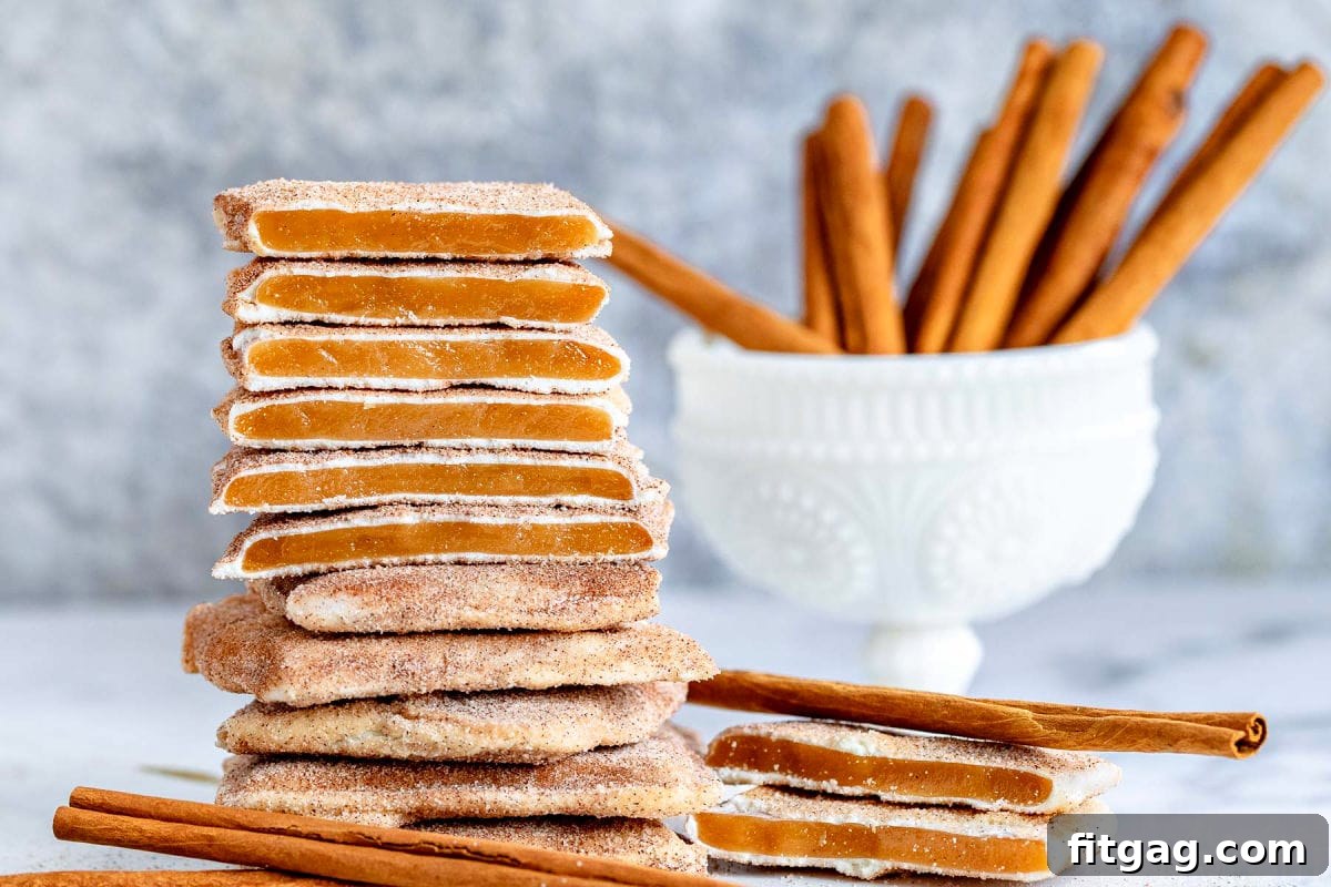 Square churro toffee stacked, showing the toffee, white chocolate, and cinnamon sugar layers with cinnamon sticks in the background.