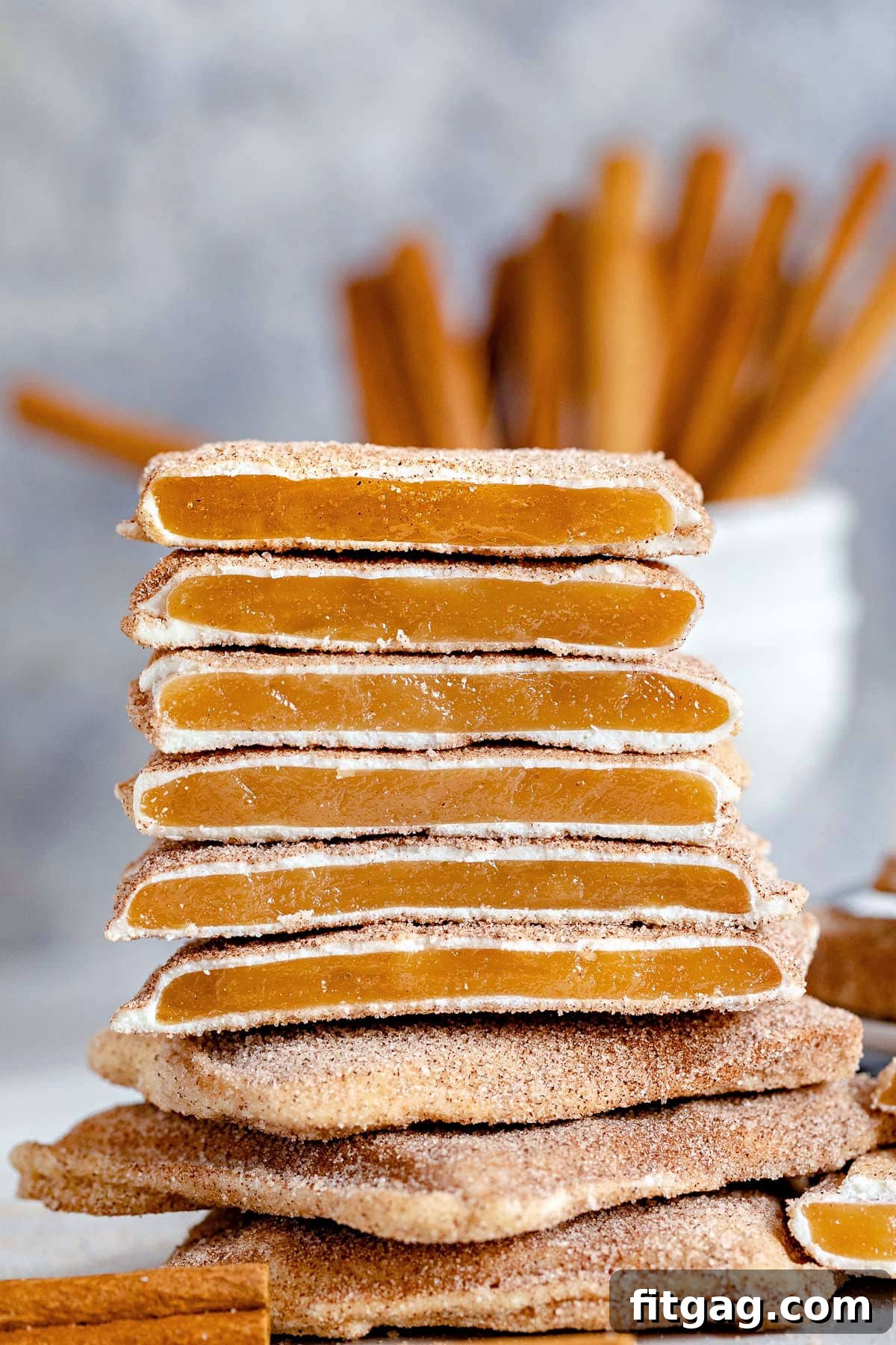 Square churro toffee stacked, showcasing the toffee, white chocolate, and cinnamon sugar layers with cinnamon sticks in the background.