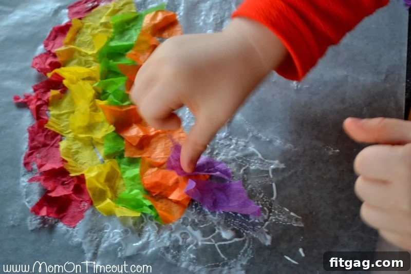 Sparkling Rainbow Suncatchers 5 Child's hands placing vibrant torn tissue paper pieces onto a glue-covered waxed paper, creating a unique stained glass rainbow pattern.