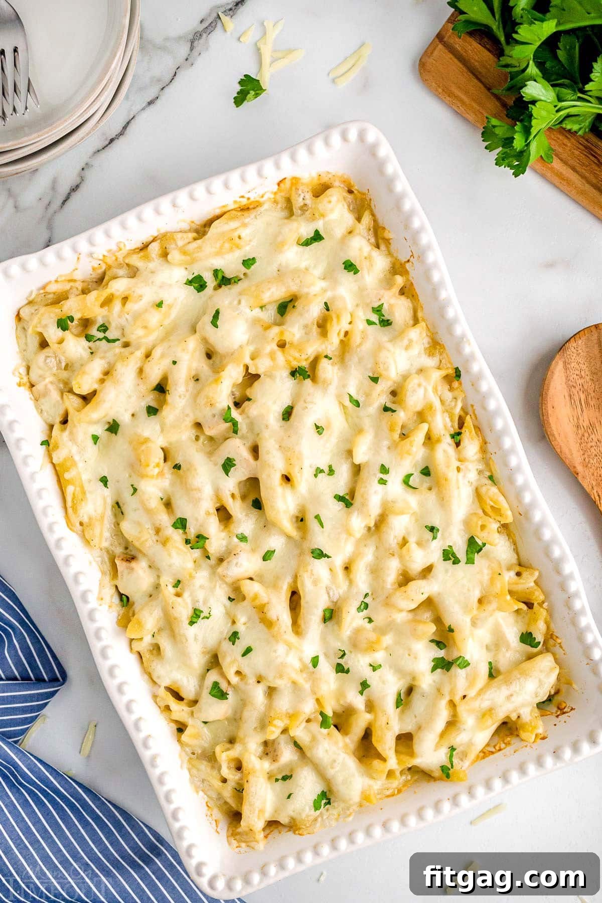 Top-down view of a golden-brown Chicken Alfredo Bake in a white casserole dish, garnished with fresh parsley and a blue and white striped napkin in the bottom left corner.