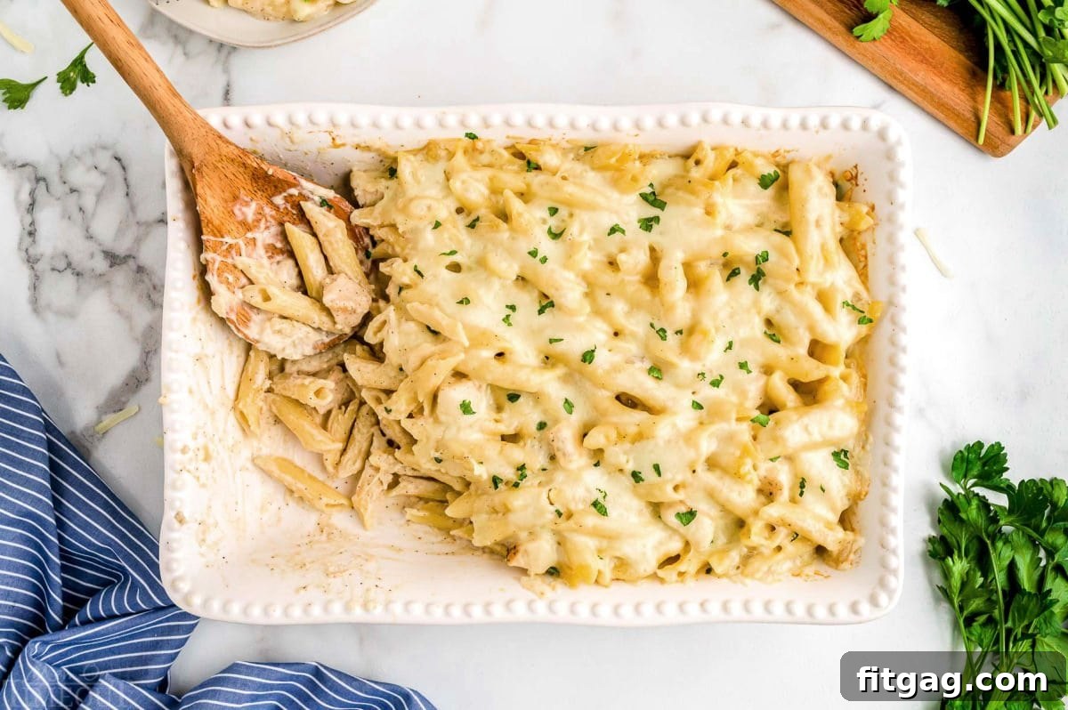 Top-down view of a white casserole baking dish with a portion of the chicken alfredo bake already served, a wooden spoon resting in the dish.