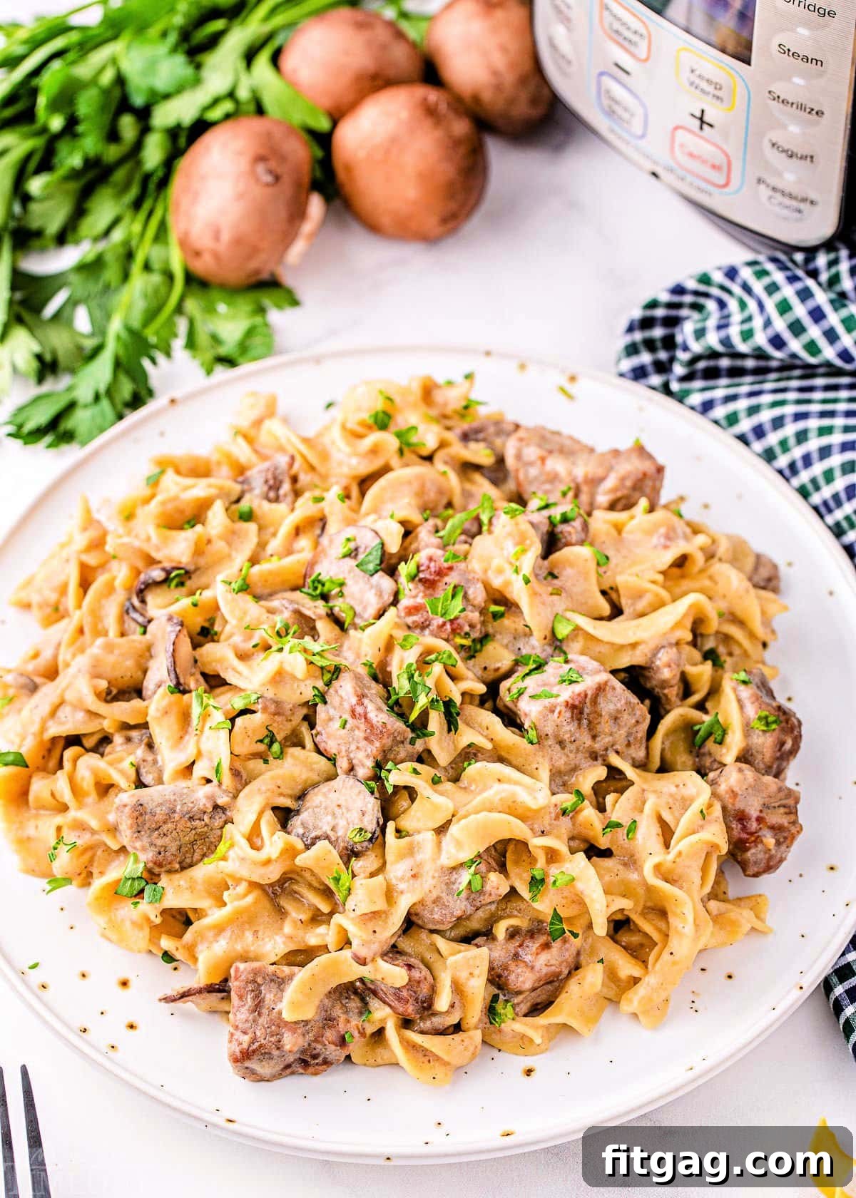 Top down look at a white plate filled with creamy Instant Pot Beef Stroganoff ready to be enjoyed. Fresh parsley and sliced mushrooms are arranged neatly in the background next to the Instant Pot.