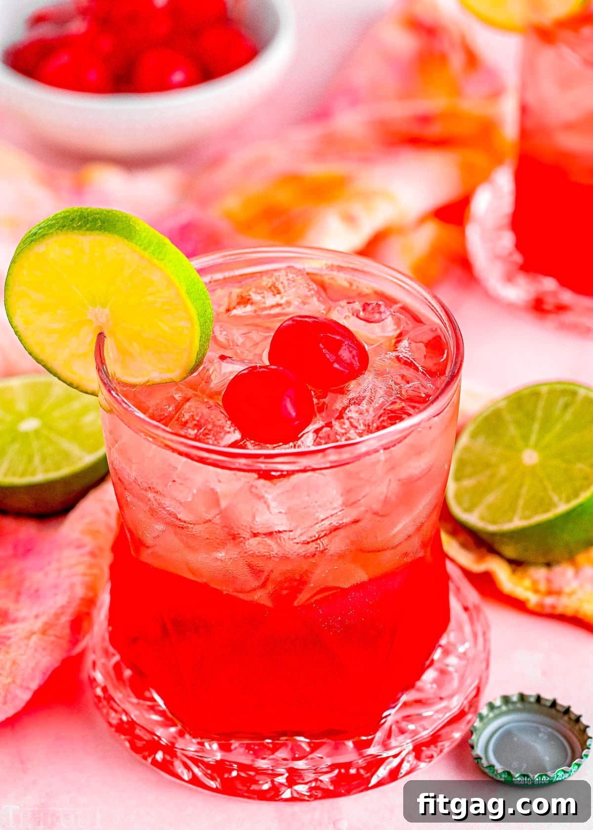 dirty shirley temple cocktail in a short glass garnished with a slice of lime and two maraschino cherries. Another drink and additional garnishes can be seen in the background.