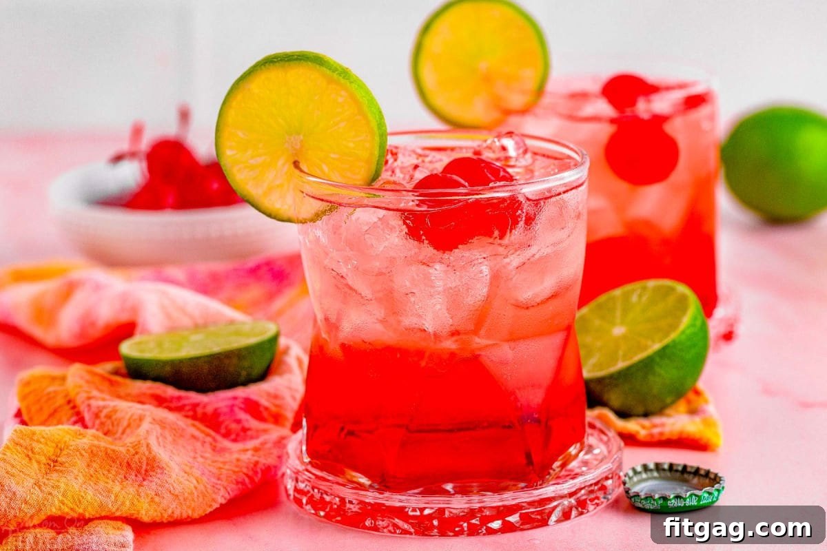dirty shirley drinks in two short glasses. garnished with a lime slice on the rim and maraschino cherries in the drinks, additional garnishes sitting on the pink marble surface around the drinks.