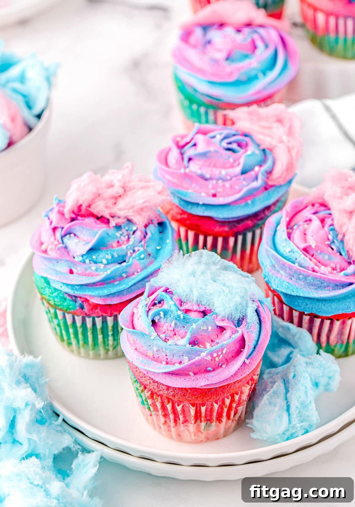 Five cotton candy cupcakes sitting on a white plate, each topped with swirled pink and blue frosting and small pieces of cotton candy. Sprinkles are visible on the frosting.