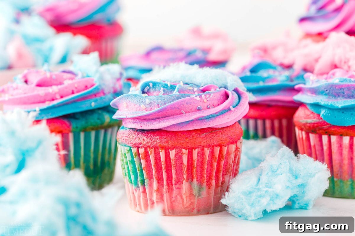A vibrant display of cotton candy cupcakes, decorated with pink and blue swirled frosting and topped with fluffy cotton candy, ready to be enjoyed. Scattered pieces of cotton candy surround the cupcakes.