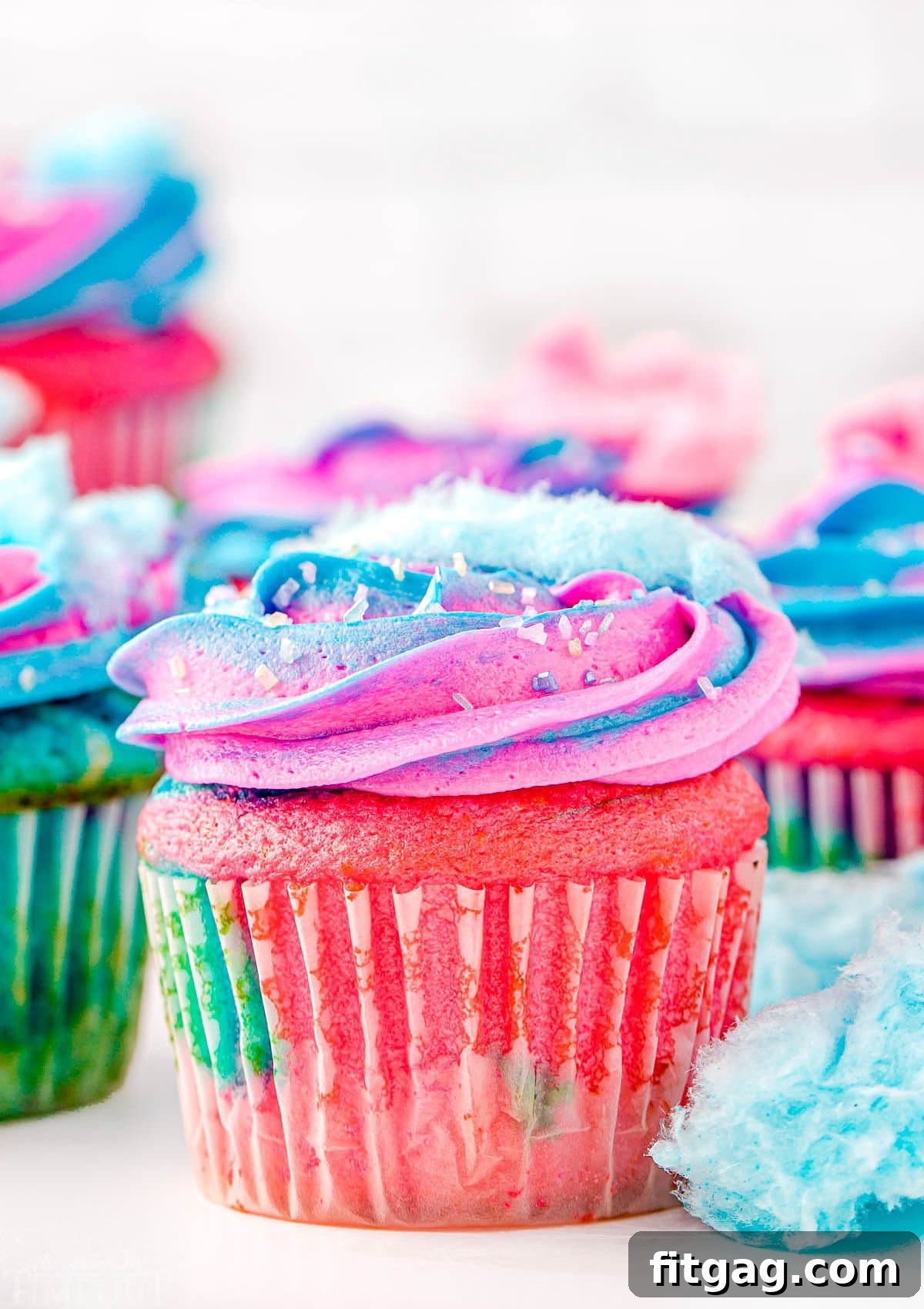 A charming arrangement of cotton candy cupcakes, beautifully decorated with pink and blue frosting swirls and topped with fluffy cotton candy. Additional cotton candy pieces are artfully scattered around the desserts.