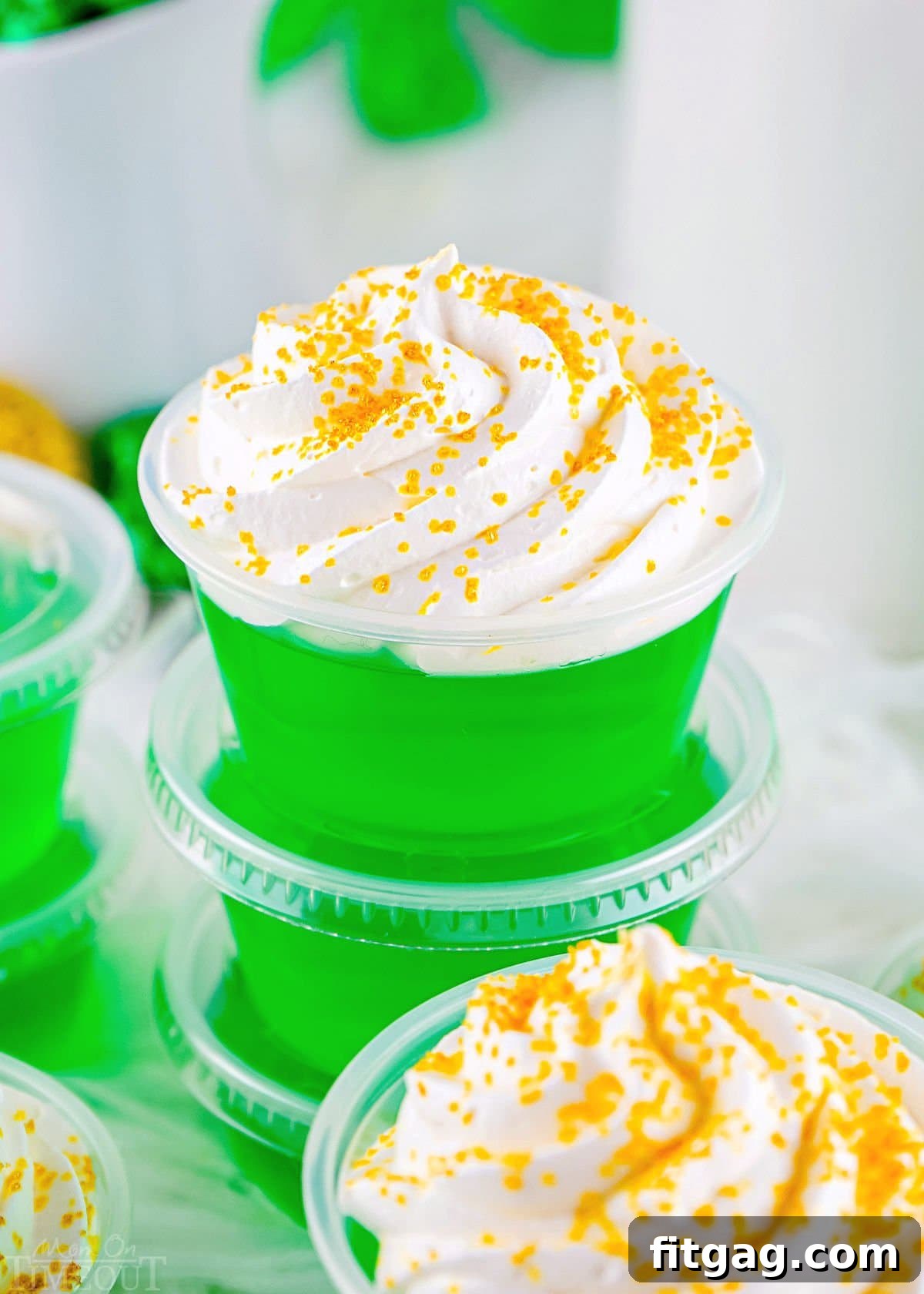 Six green jello shots in small plastic cups topped with fluffy whipped cream and shimmering gold sanding sugar, neatly stacked for a party.