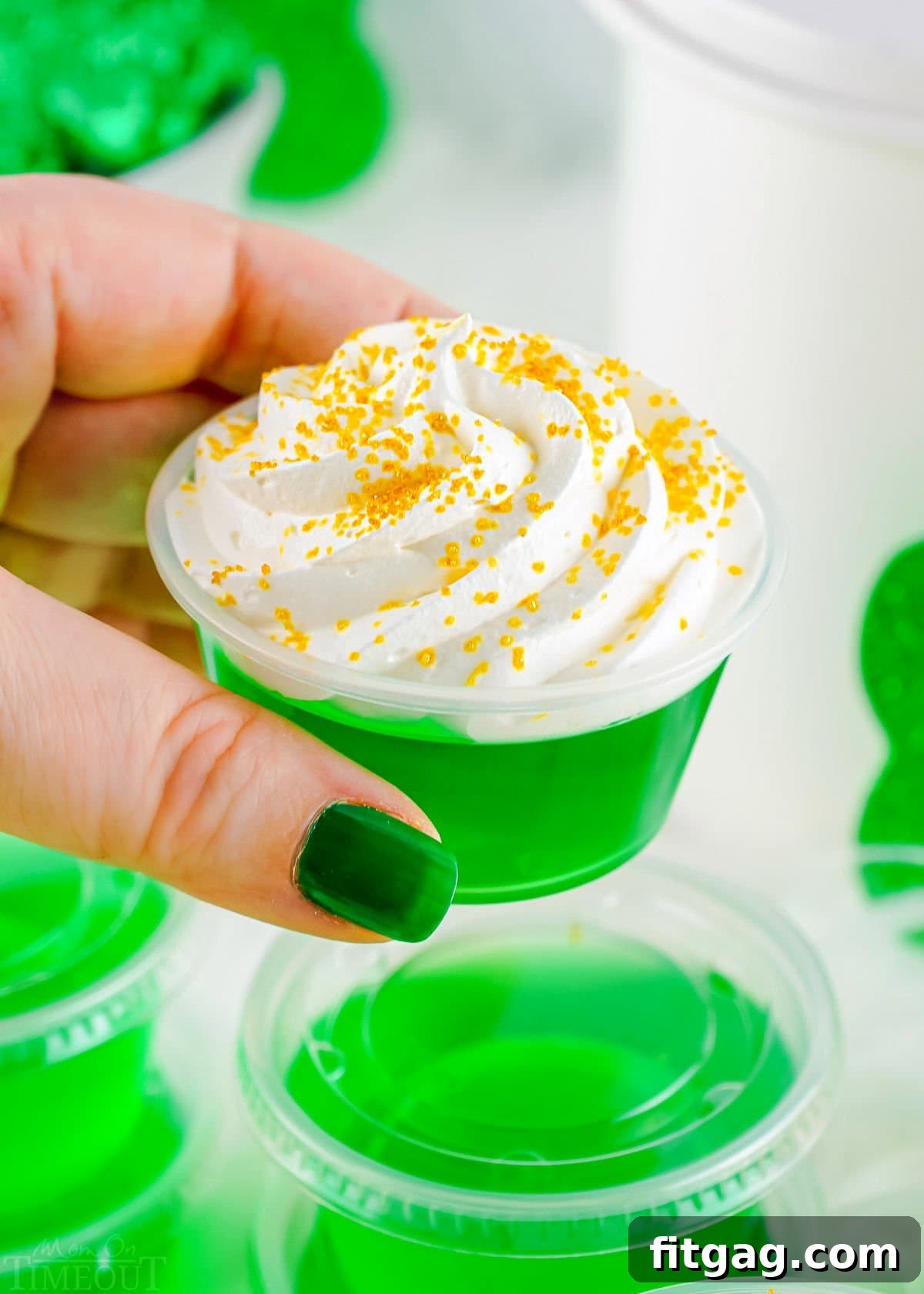 A hand holds a single green jello shot, topped with whipped cream, against a backdrop of more festive jello shots, ready for a celebration.