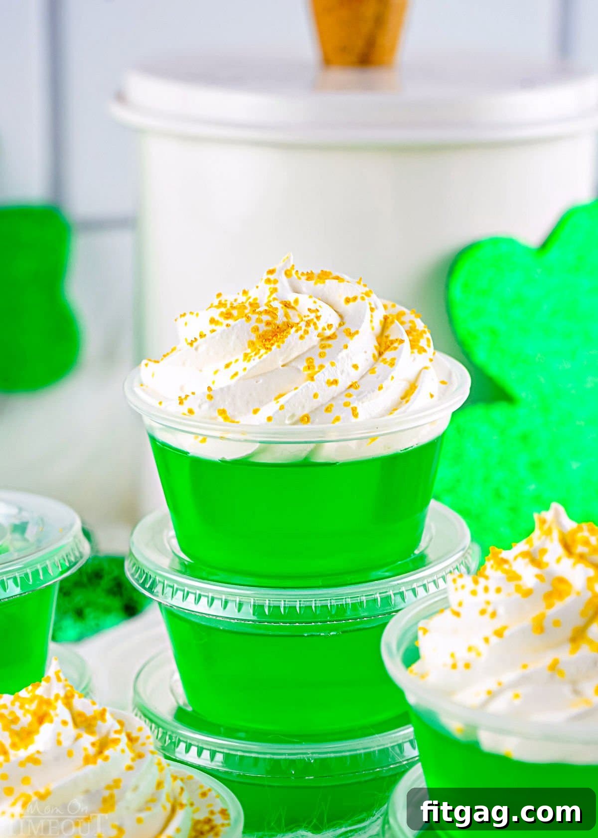 A festive arrangement of four green jello shots in small plastic cups, garnished with whipped cream and gold sanding sugar, stacked beside St. Patrick's Day decorations.
