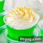Close-up of a vibrant green jello shot made with vodka, elegantly topped with fluffy whipped cream and sparkling gold sanding sugar.