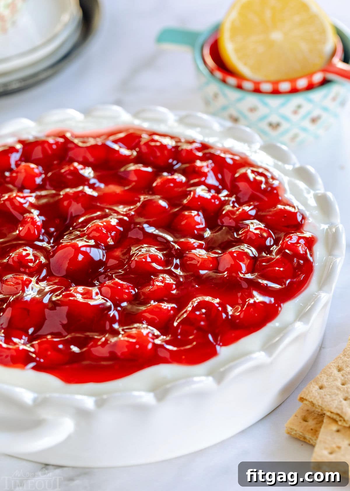 A beautiful white scalloped pie plate filled with creamy cherry cheesecake dip, generously topped with bright red cherry pie filling, ready for serving.