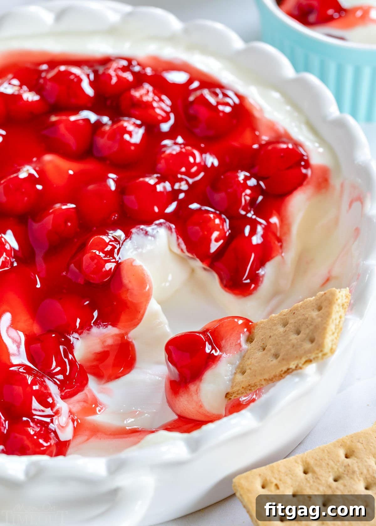 A close-up shot of creamy cherry cheesecake dip in a white pie plate, with a graham cracker dipping into the smooth, delicious mixture. A portion of the dip has already been enjoyed.