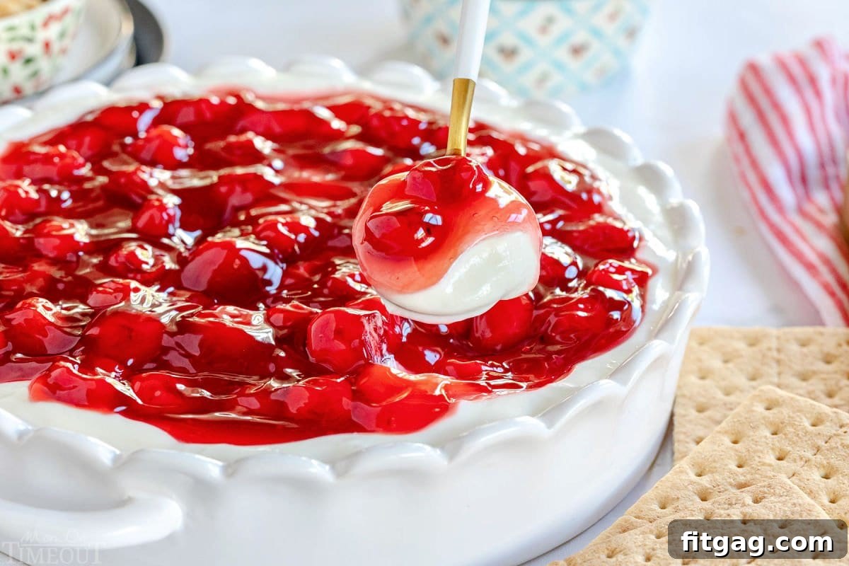 A spoon lifts a generous scoop of cherry cheesecake dip, showcasing its creamy texture and rich cherry topping, against the backdrop of the full serving dish.