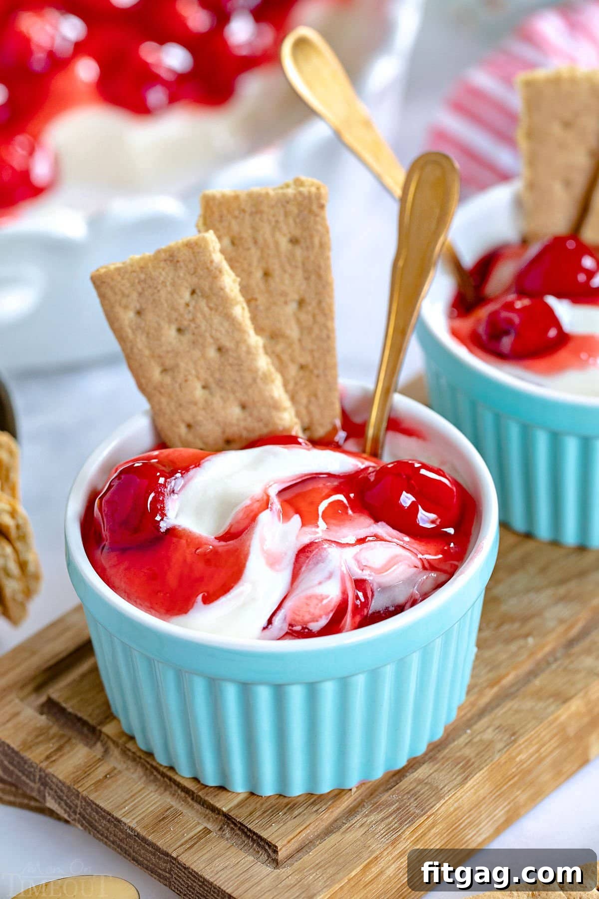 A small, elegant aqua-colored ramekin filled with individual serving of cherry cheesecake dip, topped with cherry pie filling and garnished with two graham cracker pieces and a tiny spoon.
