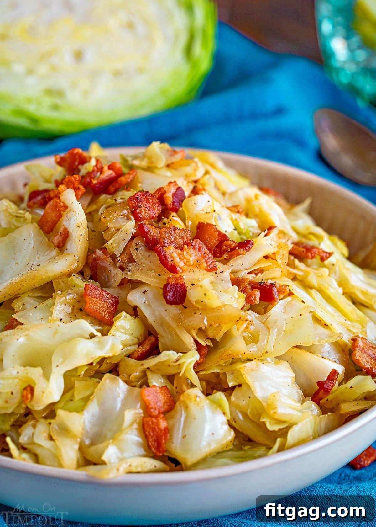 Golden Fried Cabbage 2 A shallow bowl brimming with golden-fried cabbage, perfectly mixed with crispy bacon, translucent onions, and fragrant garlic. The bowl rests on a vibrant teal napkin, with a fresh half-head of cabbage in the soft-focus background, highlighting the dish's fresh ingredients.