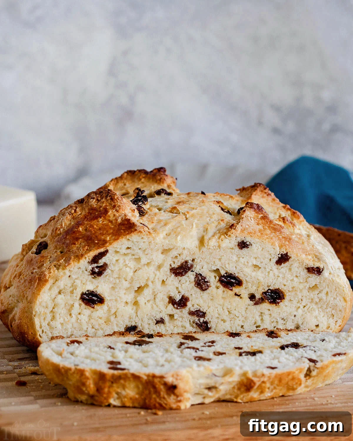 A beautifully baked loaf of Irish Soda Bread with a slice cut and placed on a wooden cutting board, with a soft blue napkin in the background, highlighting its rustic appeal.