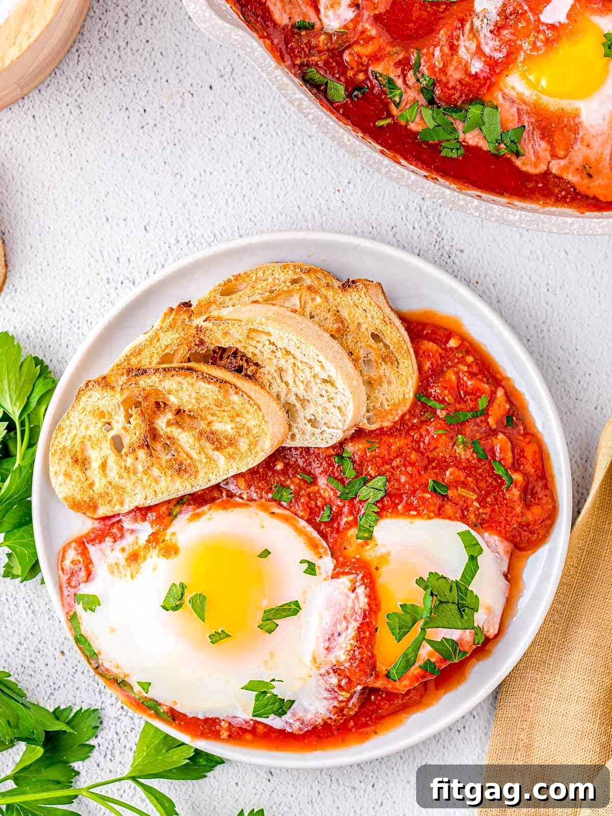 Top-down view of a plated Eggs in Purgatory dish, showcasing two perfectly poached eggs resting in a vibrant red tomato sauce. A slice of crusty, toasted bread is on the side, and the dish is garnished with fresh chopped parsley.