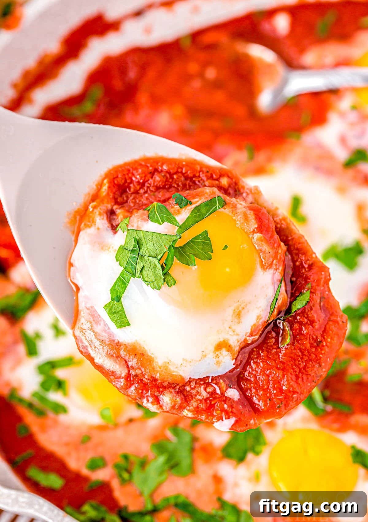 Close-up of a spoon held over a skillet, lifting a perfectly poached egg and a scoop of rich tomato sauce.