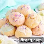 wide view of colorful italian cookies piled high on a white scalloped plate. cookies are topped with sprinkles and pastel glaze in varying colors.