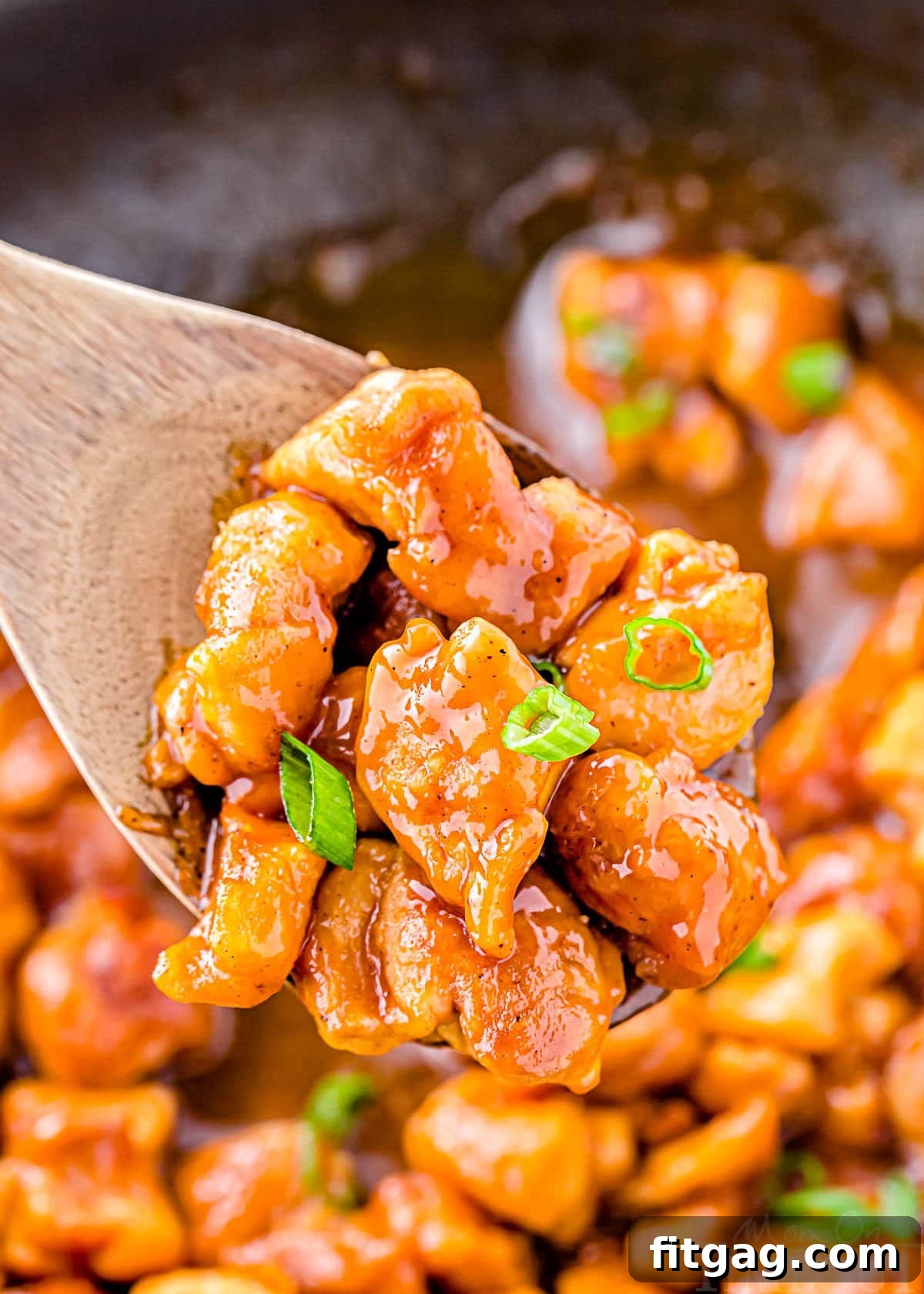 Sticky Honey Garlic Chicken 4 wooden spoon held above skillet with honey garlic chicken. garnished with sliced green onions.
