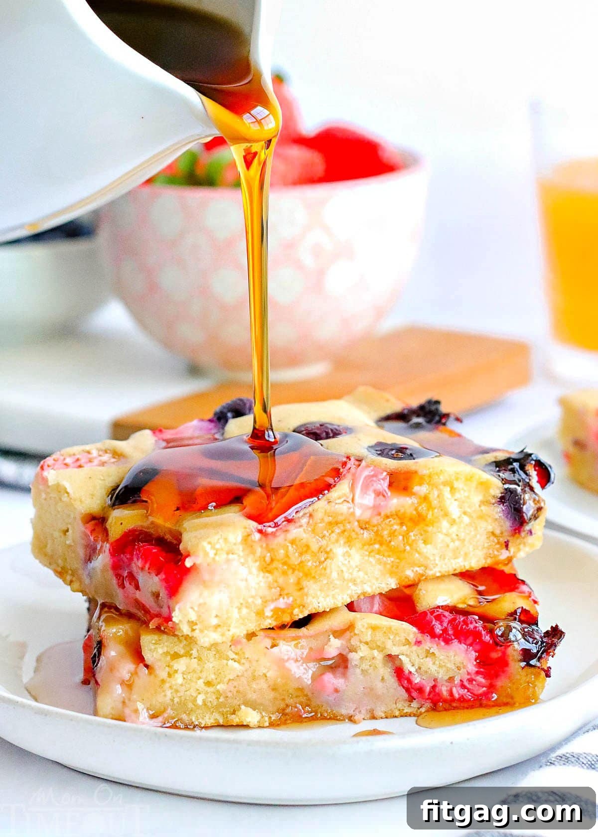 Oven Pancakes 2 Two generous pieces of sheet pan pancakes, generously topped with a vibrant mix of fresh strawberries, blueberries, and raspberries, are elegantly stacked on a pristine white plate. A stream of golden syrup cascades over the pancakes, while a bowl brimming with fresh berries adds a delightful touch to the background.
